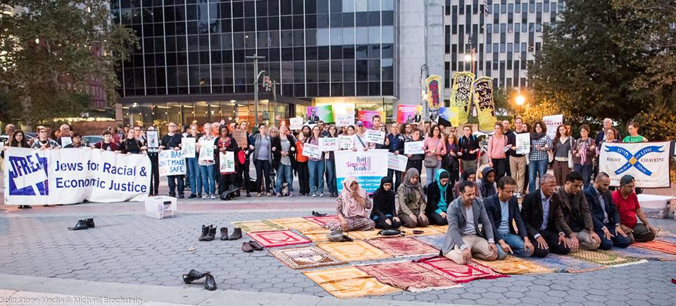 Jews and Muslims gather outside, some on prayer rugs and others behind holding protest signs.